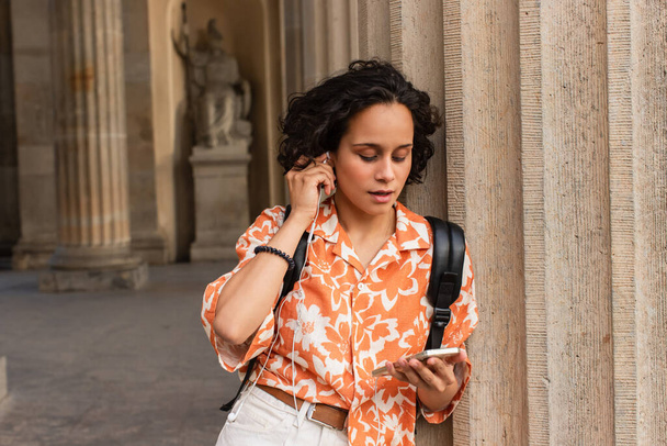 young woman adjusting wired earphones while using smartphone near blurred statue in berlin  - Photo, Image