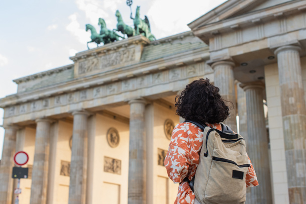 back view of curly tourist with backpack standing near brandenburg gate in berlin  - Photo, Image