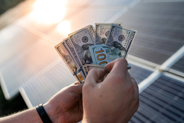 Manos masculinas con billetes de dólar contra paneles solares. Concepto de ahorro - Foto, Imagen