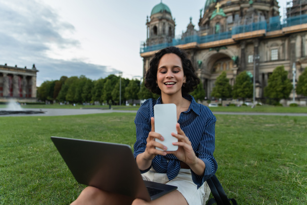счастливая женщина сидит с ноутбуком и делает селфи на смартфоне возле собора в Берлине - Фото, изображение