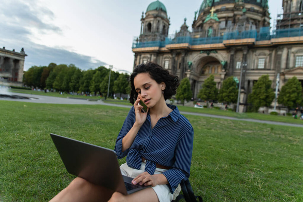 σγουρή νεαρή γυναίκα κάθεται με φορητό υπολογιστή και μιλάμε σε smartphone κοντά στον καθεδρικό ναό στο Βερολίνο - Φωτογραφία, εικόνα