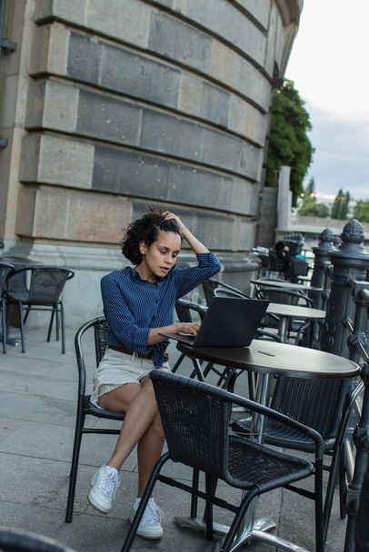 young freelancer adjusting curly hair and using laptop while sitting on summer terrace in berlin  - Foto, imagen
