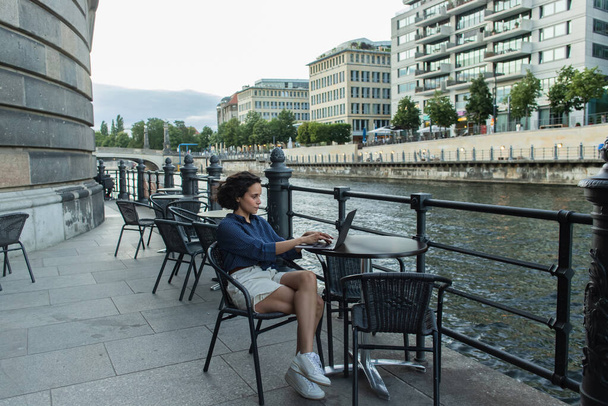полный рост молодого фрилансера, использующего ноутбук, сидя на летней террасе у реки в Берлине  - Фото, изображение
