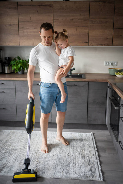 Father with his daughter on his hands vaccuming the floor in the modern home kitchen. High quality photo - Photo, image