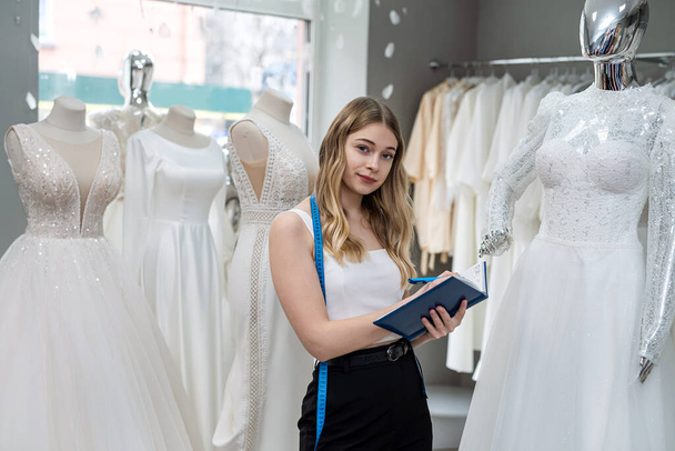 Pretty  female dressmaker with diary and pen posing near bridal wear in wedding fashion boutique - Fotografie, Obrázek