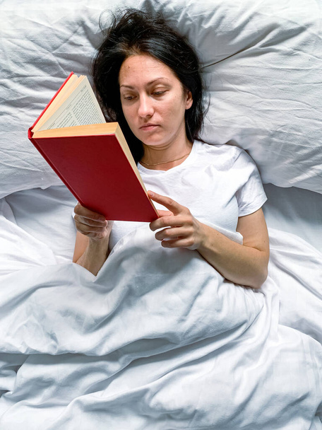 Молодая открытая женщина тысячелетия читает дома красную книгу на белых полосатых простынях. Рано утром. Текст в книге размыт. Образование или медленный образ жизни. Молодая красивая женщина - Фото, изображение