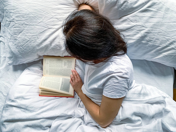 Young millennial candid woman is reading red book at home on white striped bedsheets. Early morning. Text in book is blurred. Education or slow living concept. Young adult beautiful woman - Foto, Bild