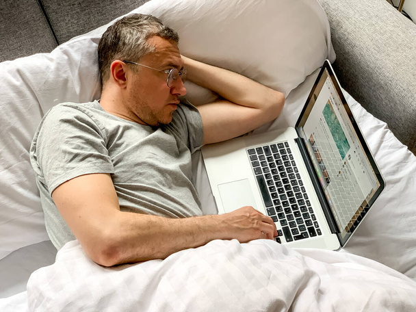 Middle aged adult man using laptop at home In bed vibes white bedsheets Alone Early morning working. freelancer, freelance job, using technology, using social media, watching movie, checking news - Zdjęcie, obraz