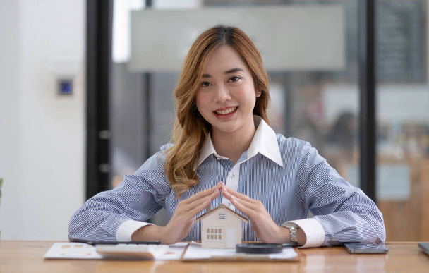 Miniature house in the hands of an Asian woman real estate agent home loan working at the office. Looking at the camera - Zdjęcie, obraz