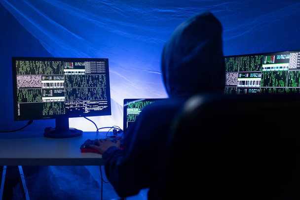 A hátsó nézet od csuklyás hacker számítógéppel a sötét szobában éjszaka, cyberwar koncepció. - Fotó, kép
