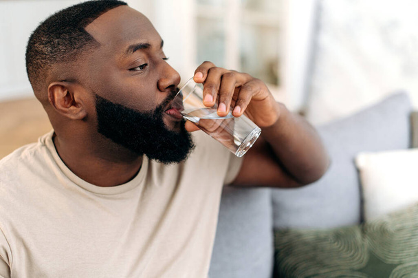 Close-up of african american man, drinks a glass of clean water with pleasure, care his health, balances fluids in the body, replenishes energy and strength in the body. Leading a healthy lifestyle - Photo, Image