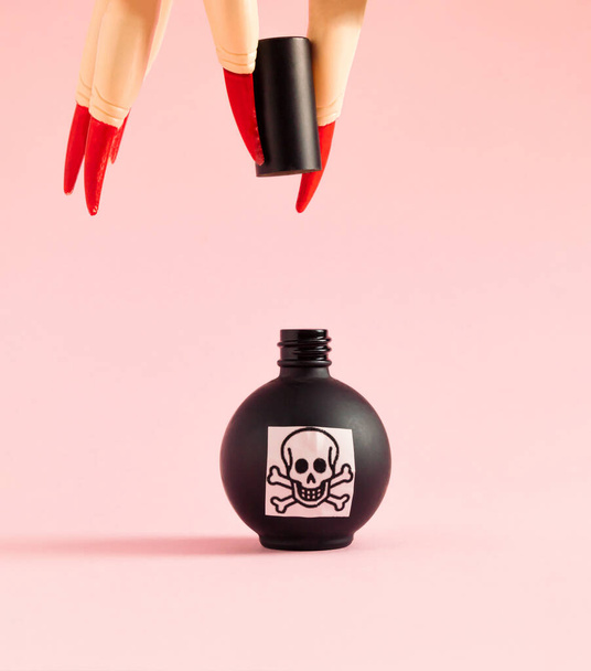 Witch's hand with red nails and poison in black bottle against pastel pink background. Halloween surreal spooky idea. - Photo, image