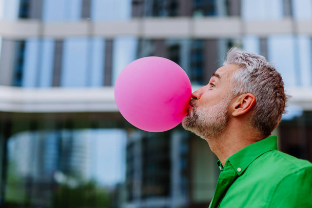 Fun portrait of happy energetic mature man inflating pink balloon in street, standing in front of a skyscraper, copy space. - 写真・画像