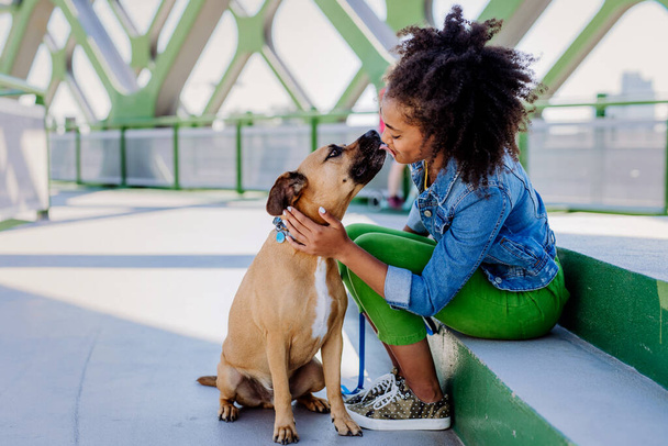 Multiracial girl sitting and resting with her dog outside in the bridge, training him, spending leisure time together. Concept of relationship between a dog and teenager, everyday life with pet. - Foto, afbeelding