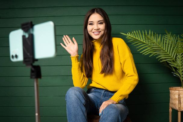 Hi, hello. Cheerful asian woman blogger greets his viewers with hand up. Girl wearing orange sweater and jeans, sitting on a green wall background and smiling to smartphone camera. - Fotó, kép