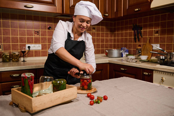 Απολαυστική πολυεθνική νεαρή γυναίκα, νοικοκυρά φορώντας λευκό σκούφο σεφ και μαύρη ποδιά, χρησιμοποιώντας ράφτη, κλείνει καπάκια βάζων, ενώ κονσερβοποιώντας ντοματίνια στην κουζίνα του σπιτιού. Πίκλες, μαρινάρισμα - Φωτογραφία, εικόνα