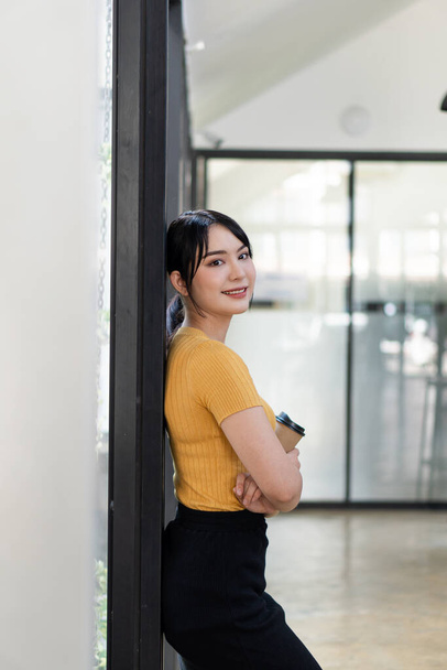 Beautiful Asian woman standing with her arms crossed with a coffee cup beside the flap in the office during work breaks. - 写真・画像