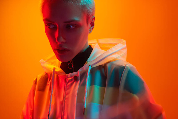 Young female model in translucent futuristic coat with short hair looking away under neon illumination against orange background - Foto, imagen