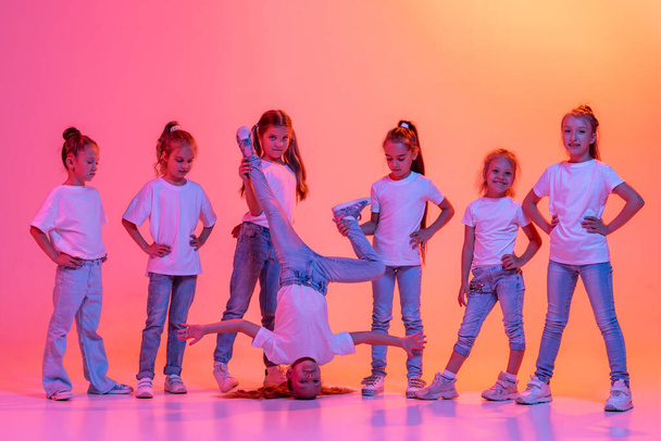 Hip-hop dance, street style. Group of children, school age girls in casual style clothes dancing in choreography class isolated on pink background in yellow neon light. Concept of music, fashion, art - Photo, Image