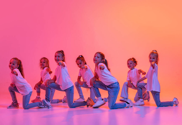 Modern contemp dance. Happy kids, school age girls in white t-shirts and jeans dancing in choreography class isolated on pink background in yellow neon light. Concept of music, fashion, art - Photo, Image