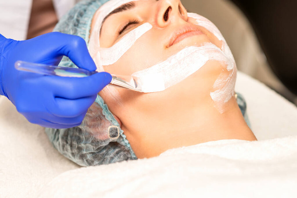 Ο αισθητικός με πινέλο εφαρμόζει μια φωτοχημική και γλυκολική μάσκα peeling προσώπου στο γυναικείο πρόσωπο ασθενούς στην κλινική ομορφιάς - Φωτογραφία, εικόνα
