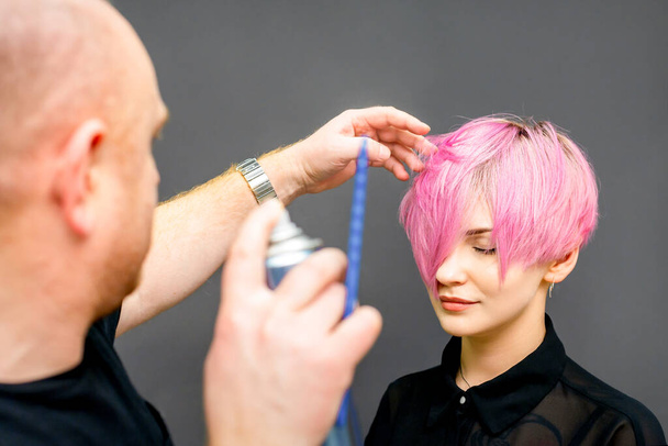 The hairdresser is using hair spray to fix the short pink hairstyle of the young caucasian woman in the hair salon - Foto, Imagen