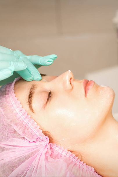 Cosmetologo con le mani guantate applica una maschera idratante con crema peeling sul viso femminile. Trattamento cosmetologico facciale. Procedure per la cura del viso - Foto, immagini