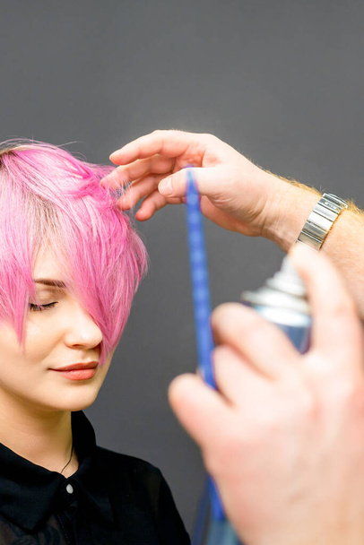 The hairdresser is using hair spray to fix the short pink hairstyle of the young caucasian woman in the hair salon - 写真・画像