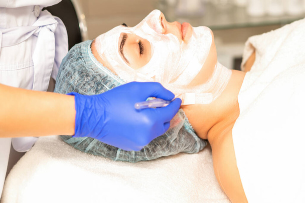 Ο αισθητικός με πινέλο εφαρμόζει μια φωτοχημική και γλυκολική μάσκα peeling προσώπου στο γυναικείο πρόσωπο ασθενούς στην κλινική ομορφιάς - Φωτογραφία, εικόνα