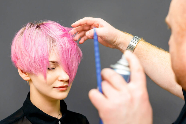 Парикмахер использует лак для волос, чтобы исправить короткую розовую прическу молодой кавказки в парикмахерской. - Фото, изображение