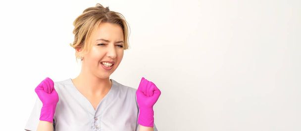 Happy caucasian woman doctor wearing pink gloves celebrates and raising fists on white background - Photo, Image