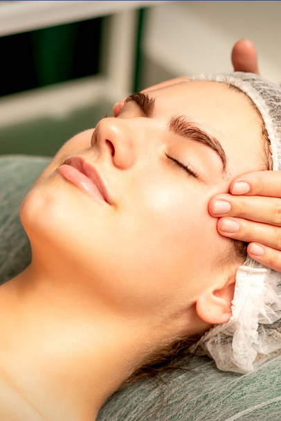 Masaje de cabeza. Hermosa mujer blanca joven caucásica recibiendo un masaje en la cabeza y la frente con los ojos cerrados en un salón de spa - Foto, imagen