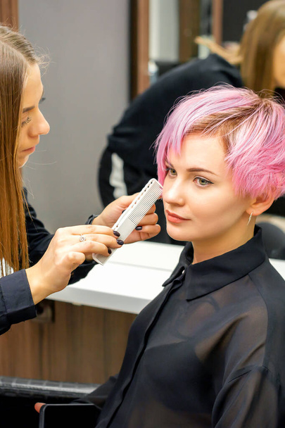 Hairdresser with comb is checking out and fixing the short pink hairstyle of the young white woman in a hair salon - Photo, image