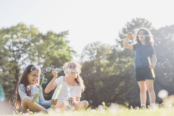 Three beautiful caucasian girls sisters are sitting on a flower lawn in a public park and blowing soap bubbles in the backlight with a blurred background, close-up side view. PARKS REC concept, happy - 写真・画像