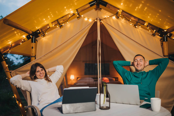 Счастливая семейная пара фрилансеров, работающих ноутбуком в уютной заклеенной палатке летним вечером. Люксовый кемпинг-шатер для отдыха на свежем воздухе. Концепция образа жизни - Фото, изображение