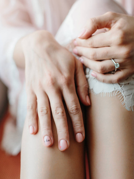 A bride with a nude manicure in an engagement ring with a gemstone keeps her hands on her knees. On the bride's ring finger on the nail, inscribe "I do" - Φωτογραφία, εικόνα