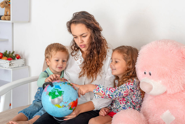Маленькие дети с няней или с молодой матерью или с учителем сидят на диване в комнате и изучают земной шар - Фото, изображение