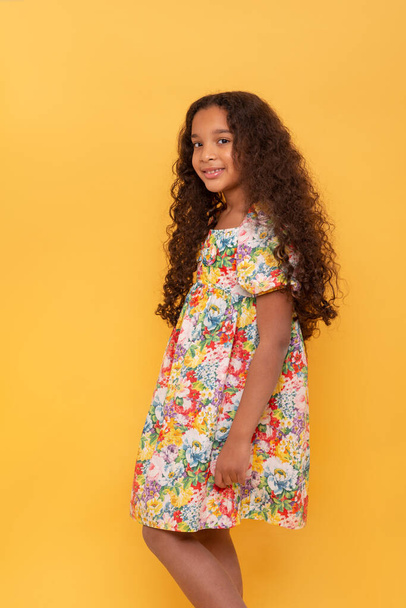 Half length portrait of a dark skinned young girl with long curly hair wearing colorfull dress against yellow background - Foto, afbeelding