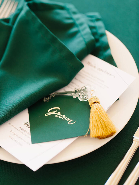 Wedding banquet table setting in emerald green. On a white plate are a restaurant menu and a folded napkin and a green sign for the groom's seat. - Valokuva, kuva