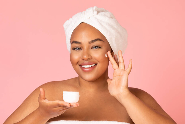 Smiling Black Female Applying Moisturizer On Face Enjoying Facial Skincare Routine Posing Over Pink Studio Background. Cosmetics For Skin And Beauty Care Concept - 写真・画像
