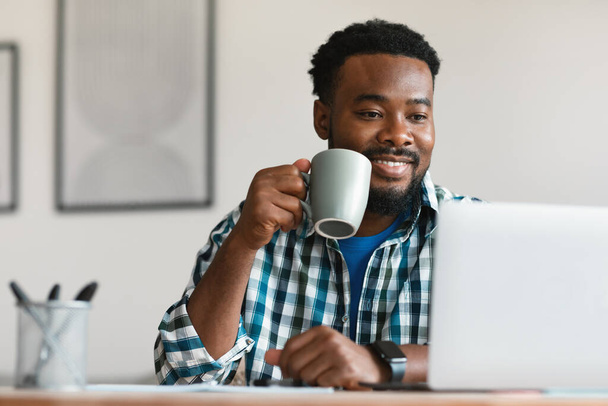 Contented Black Male Freelancer Having Coffee Using Laptop Computer Working Online Sitting At Workplace In Modern Office. Professional Business Career And Internet Technology Concept - Photo, image