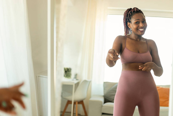 Молода темношкіра жінка в спортивному одязі танцює перед дзеркалом, маючи домашню фітнес-тренування та посміхаючись. Спортивна леді працює в приміщенні і розважається
 - Фото, зображення
