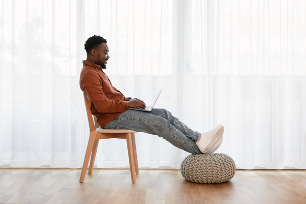 Πλευρική άποψη του μαύρου άντρα χρησιμοποιώντας Laptop εργασίας σε απευθείας σύνδεση κάθεται στην καρέκλα κοντά στο παράθυρο στο σπίτι. Επιτυχής Freelancer Guy Websurfing μέσω υπολογιστή στο Σαββατοκύριακο. Έννοια τρόπου ζωής τεχνολογίας - Φωτογραφία, εικόνα