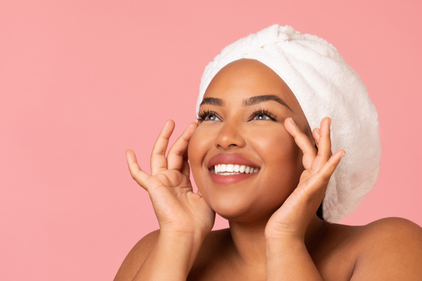 Facial Skincare. Happy African American Woman Moisturizing Face Skin Applying Moisturizer Posing With Wrapped Towel On Head Looking Aside Over Pink Background. Studio Shot - Foto, imagen