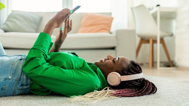 African american lady wearing wireless headphones and using smartphone, lying on floor at home, woman listening music or scrolling social networks feed, side view - 写真・画像
