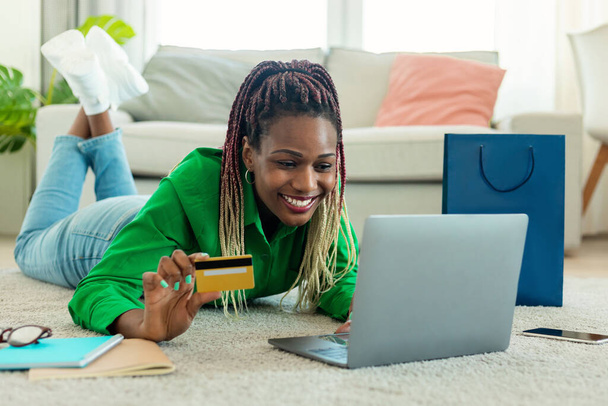 Its time for shopping. Happy african american woman using laptop and holding credit card, making purchases, buying gifts and holiday presents, lying on the floor carpet - Foto, Bild