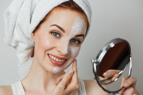 Portrait of young caucasian woman with white cosmetic mask on half of her face holding mirror against grey background. Regular beauty treatment for skin care. - Foto, immagini