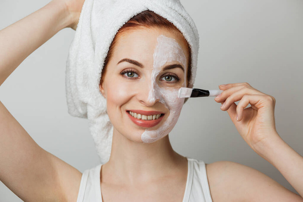Beautiful young woman in towel using brush for applying organic cosmetic mask for moisturizing facial skin. Isolated over grey studio background. Cosmetology concept. - Photo, Image