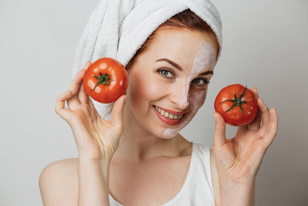 Pleasant woman in towel with cosmetic moisturizing mask on half of her face holding two fresh tomato. Charming lady with bare shoulders posing over grey background. - Φωτογραφία, εικόνα