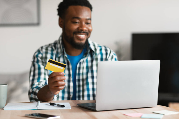 Χαμογελώντας μαύρο άντρα ψώνια σε απευθείας σύνδεση στο φορητό υπολογιστή κρατώντας πιστωτική κάρτα κάθεται στο γραφείο στο σπίτι. Ο τύπος που κάνει πληρωμές μέσω Internet Banking Service. Ecommerce and Financial Concept. Επιλογή εστίασης - Φωτογραφία, εικόνα